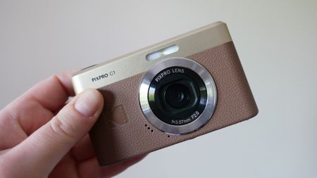 A person holding a Kodak Pixpro C1 camera with a stylish design, showcasing its lens and textured body in a well-lit setting