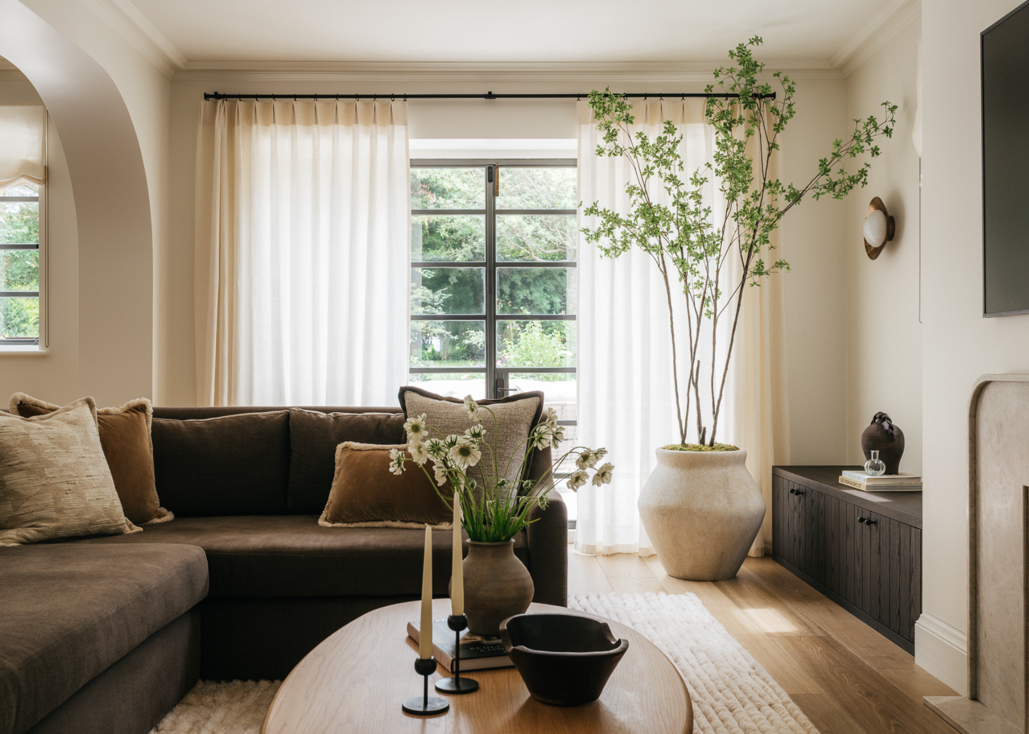 A modern living room with off-white walls, cream sheer floor-length curtains partly closed over the black-framed patio doors, and an L-shaped brown sofa in front of a coffee table with a vase of flowers, a bowl, and two taper candles; a large ceiling-height plant sits in the corner of the room by the patio doors