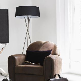 A tall tripod lamp with dark coloured shade stads behind an armchair in a white living room.