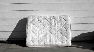 Image shows a white mattress lent against a white wooden wall outside a house on a sunny day