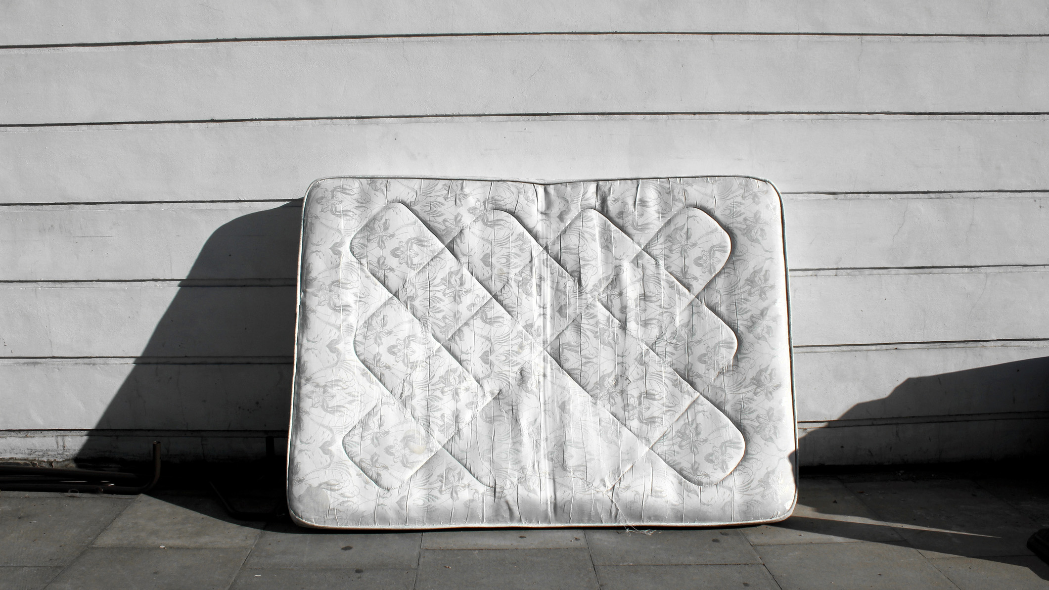 Image shows a white mattress lent against a white wooden wall outside a house on a sunny day