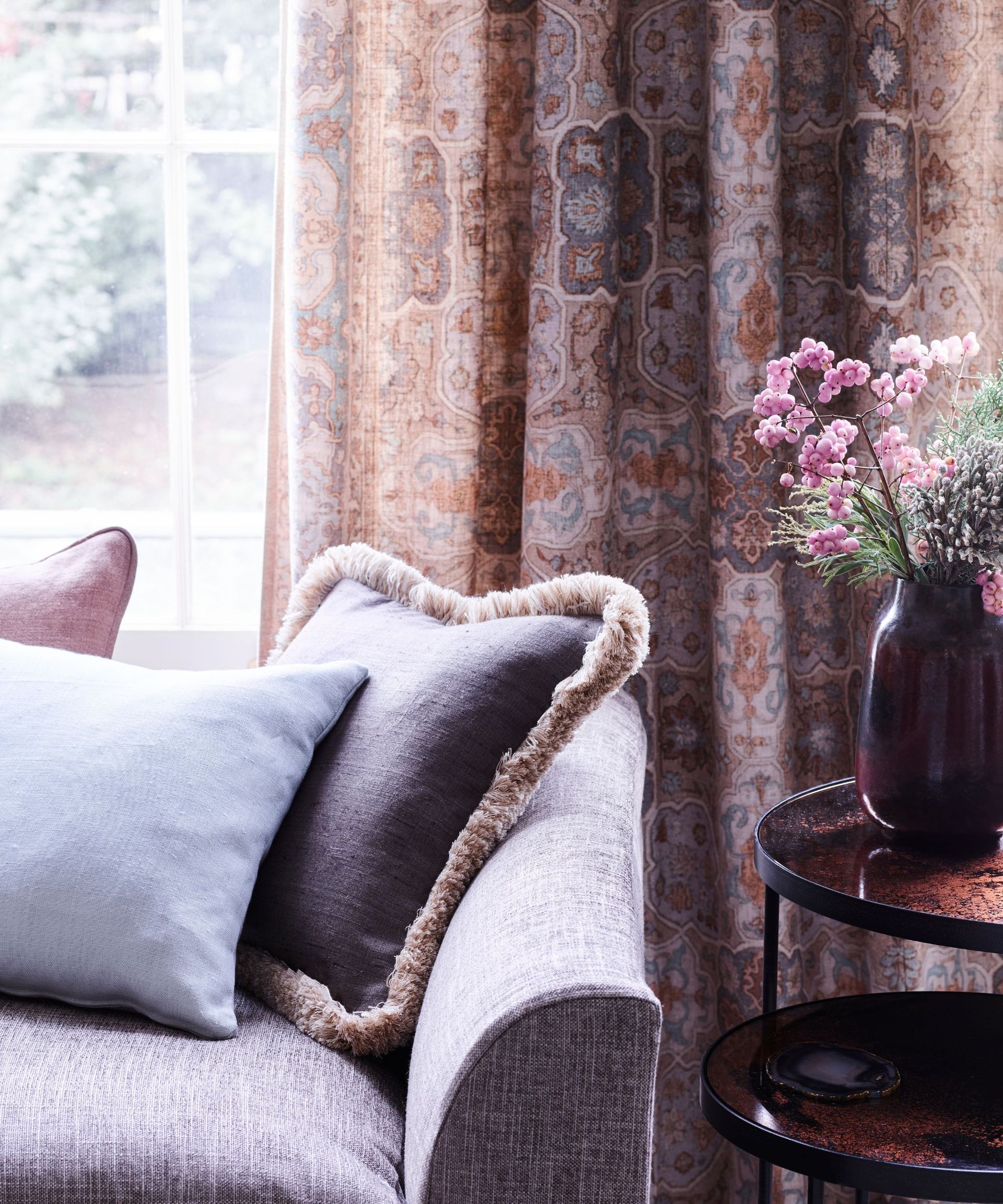 Close up of one corner of a lilac sofa with scatter cushions on in front of a window with an orange paisley printed curtain and a copper-hued side table with pink flowers in a brown vase