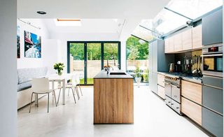 a light filled single storey kitchen extension, with black and wood kitchen counters, a wooden kitchen island in the middle, and a dining table, with a white floor, and bifold doors leading out onto a garden