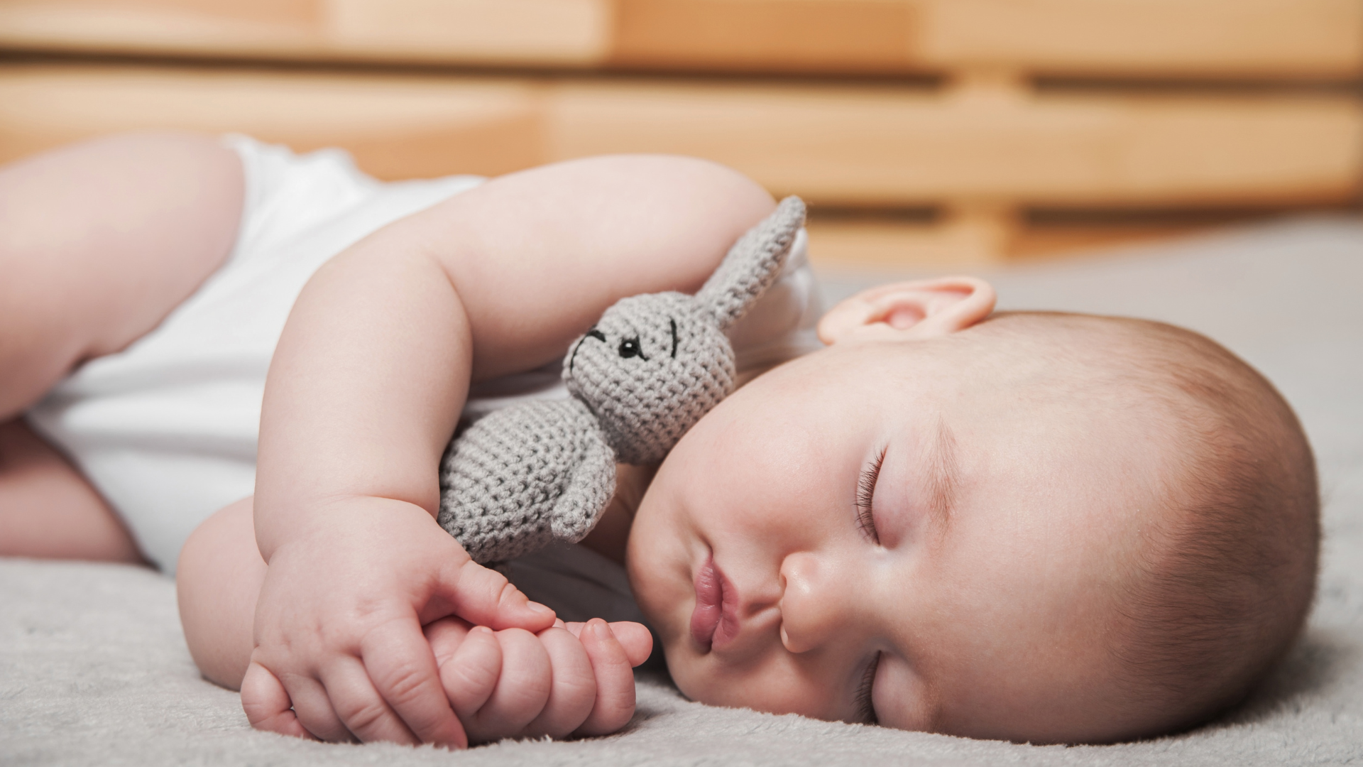 A young baby lies asleep on their side wearing a white onesie and holding a knitted gray rabbit.