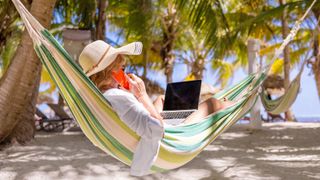 Woman lying in hammock on tropical beach and working on laptop and speaking on phone