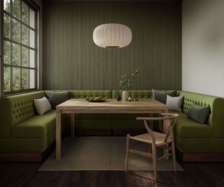 Cosy dining nook featuring deep green tufted banquette seating, a wooden dining table, matching wooden chair, and soft lighting, framed by green panelled walls and a large window.