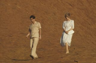 Prince Charles wearing tan pants and a shirt and Princess Diana wearing a white dress walking through a dusty landscape and looking down