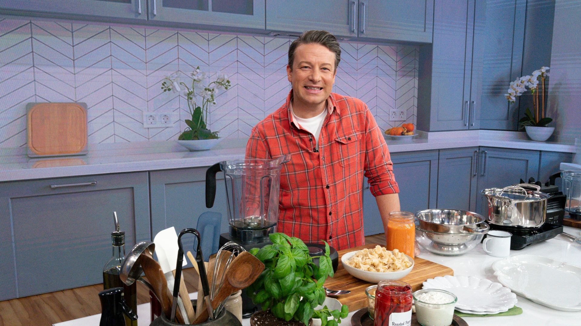 Jamie Oliver poses in a kitchen surrounded by ingredients and equipment 