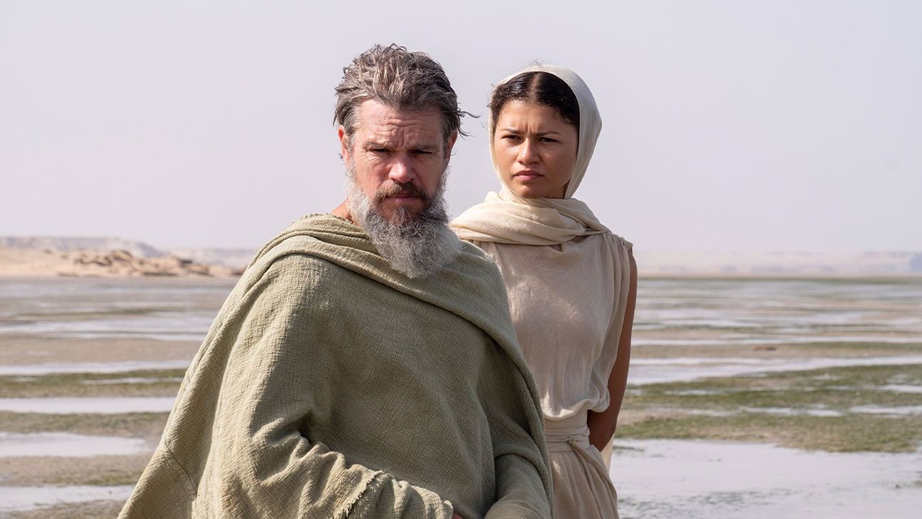 A bearded man in rags (Matt Damon as Odysseus) and a woman in a white headscarf and dress (Zendaya as Athena) stand on a beach, in 'The Odyssey.'