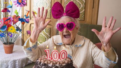 A woman wearing heart-shaped sunglasses celebrates her 100th birthday with cake and fun decorations.