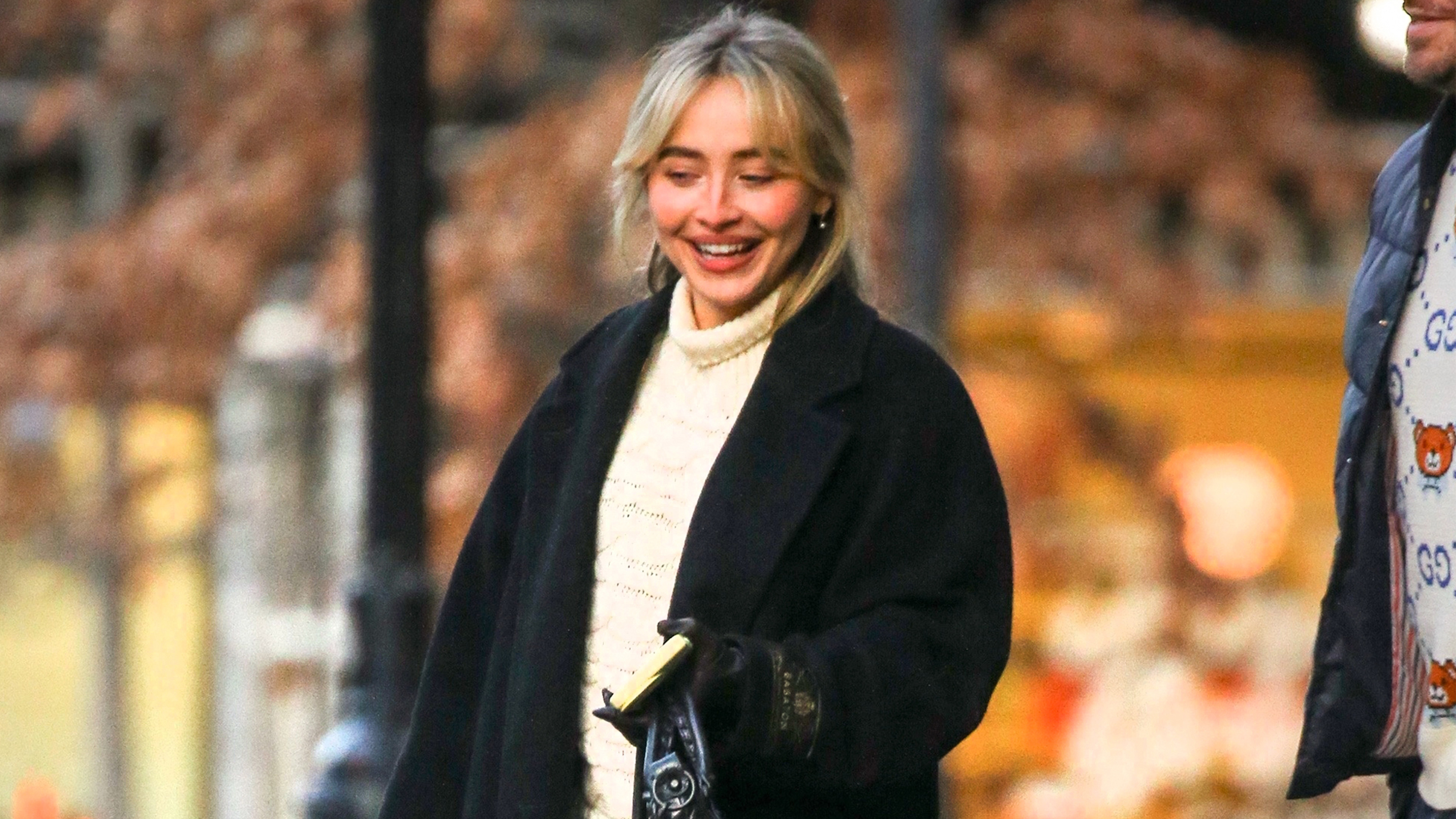 Sabrina carpenter wears an ivory turtleneck sweater, and black wool coat.