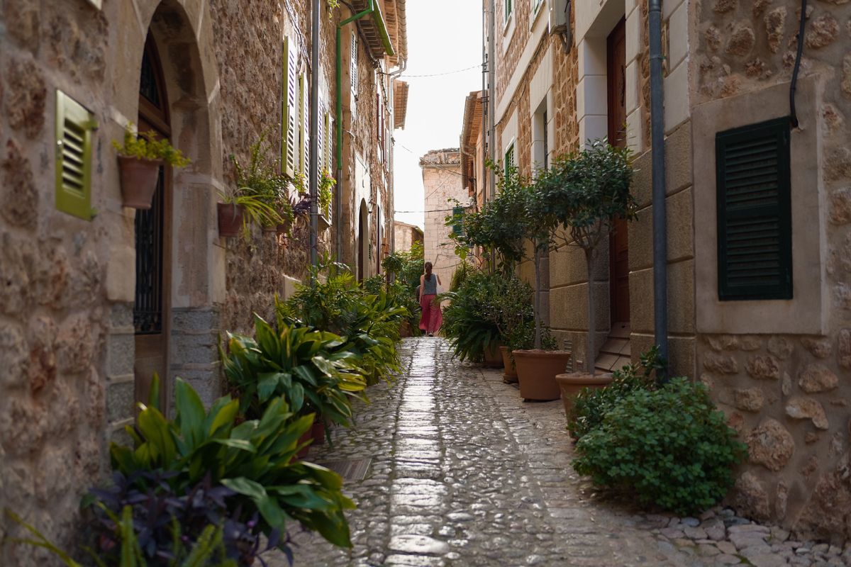 Narrow cobbled lane lined with stone houses, green shutters and potted plants, with a person walking away in the distance.