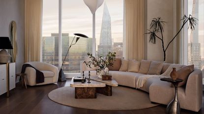 modern apartment living room with city skyline views, a curved cream couch, a cream armchair, marble nesting coffee table, round rug, and plant in corner of the room