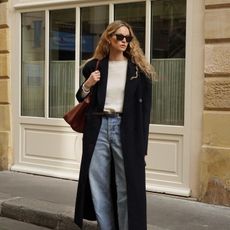 Fashion influencer Anouk Yve with long, blonde, curly hair on the street in Europe wearing belted wide-leg faded blue jeans, a cream knit sweater, a long wool coat, sunglasses, mules, and carrying a brown tote.