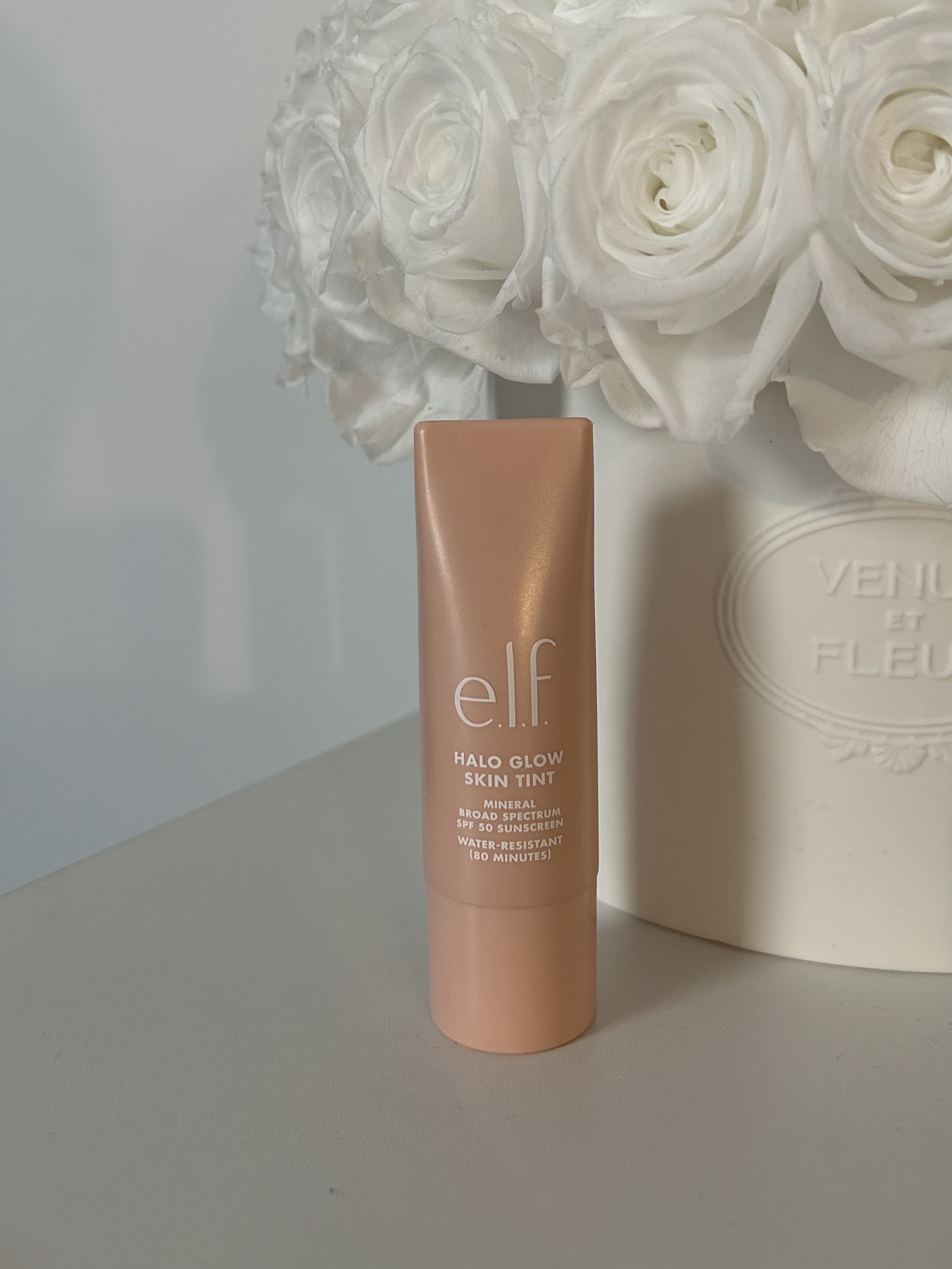 An image of the E.l.f. Cosmetics Halo Glow Skin Tint sitting in front of a vase of flowers.