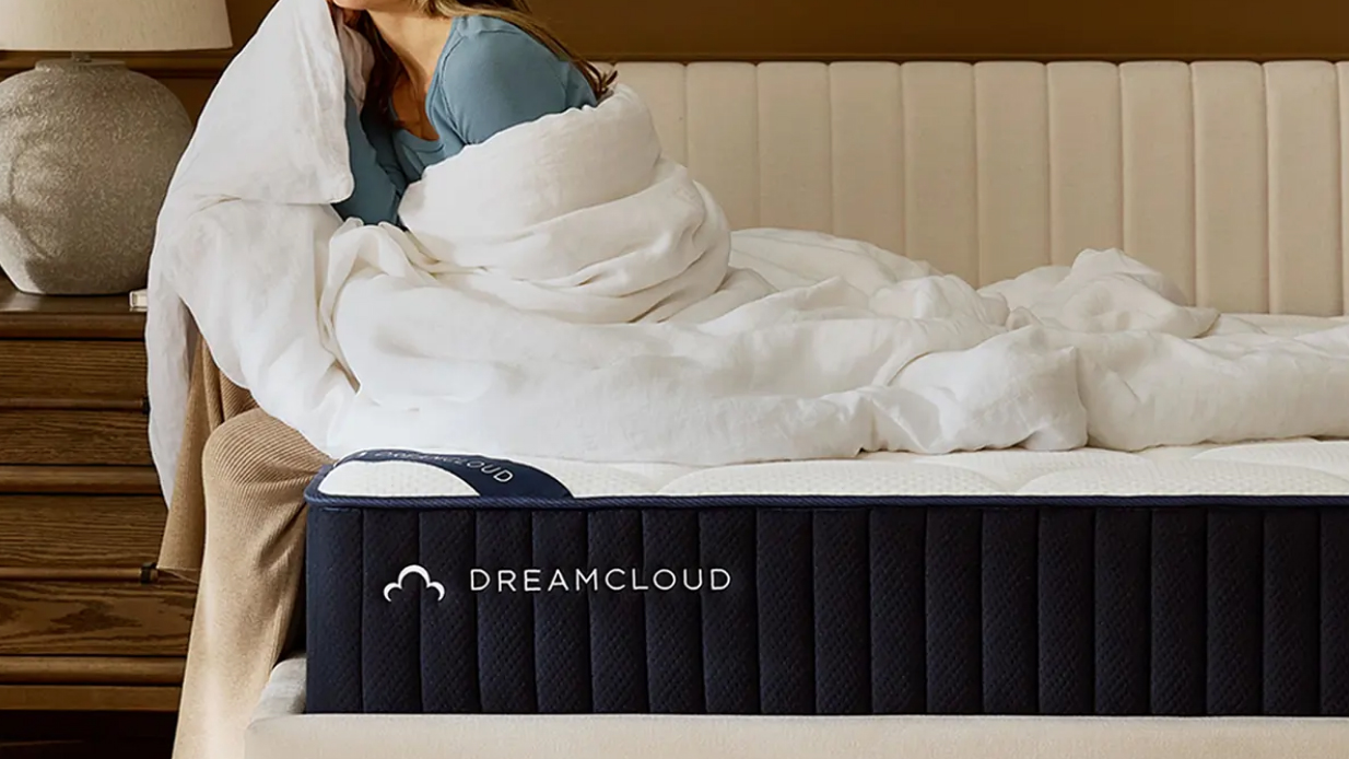 Woman sitting on the edge of the DreamCloud Classic Memory Foam Mattress wrapped in a white duvet