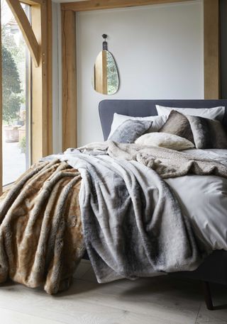 bedroom with grey bed by window, faux fur throws layered on bed with faux fur cushions/pillows, mirror on wall