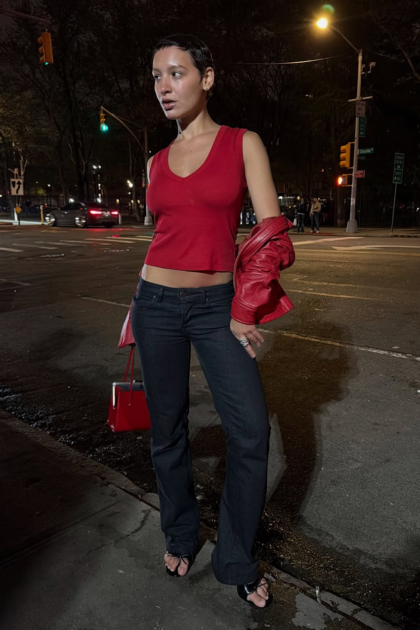 a woman wearing a red top, red leather jacket, dark wash jeans, and black sandals