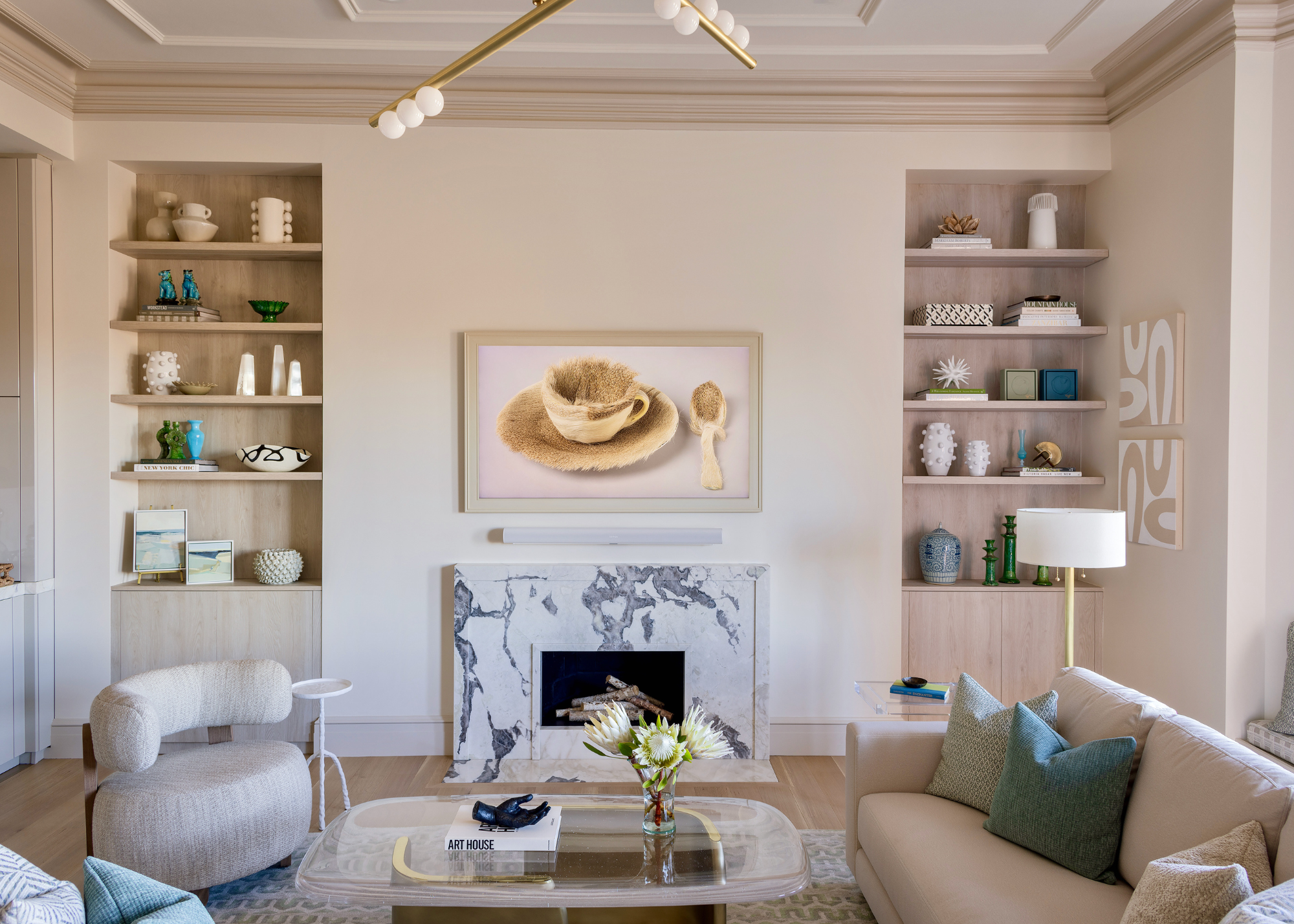 A modern neutral living room with an artwork of a fluffy coffee mug and saucer with a spoon above a marble fireplace front with alcove shelving either side filled with decorative objects