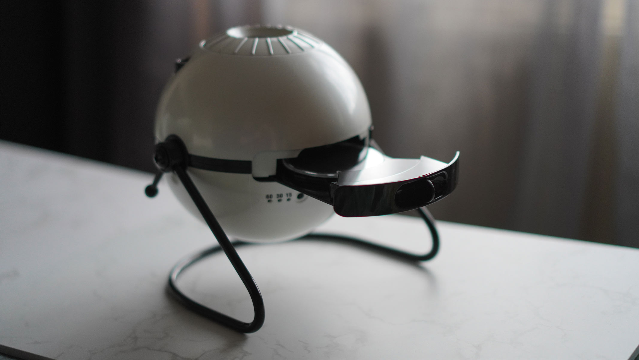 A white sphere sits on a black legged stand on top of a white surface with a black disk tray in the eject position.