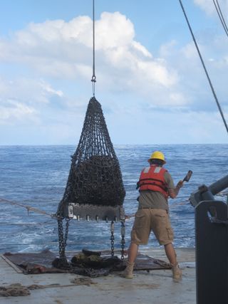 A researcher aboard a boat holds a hammer up to break apart the rocks collected from the sea floor. using a metal net.