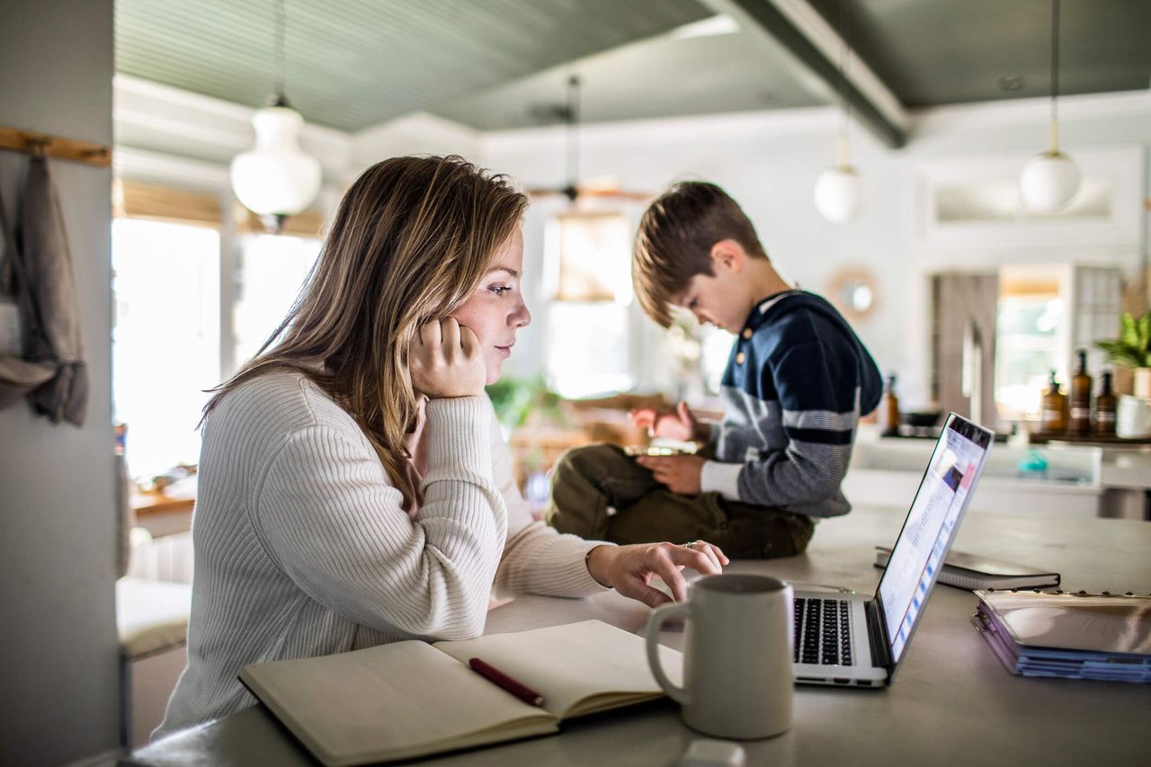 Woman working from home on laptop while son uses smartphone 