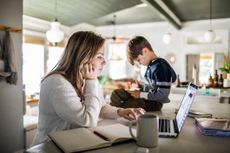 Woman working from home on laptop while son uses smartphone 