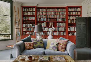 living room with bookcase painted red, grey couch, patterned pillows, grey floor, green painted window frame, ladder for bookcase