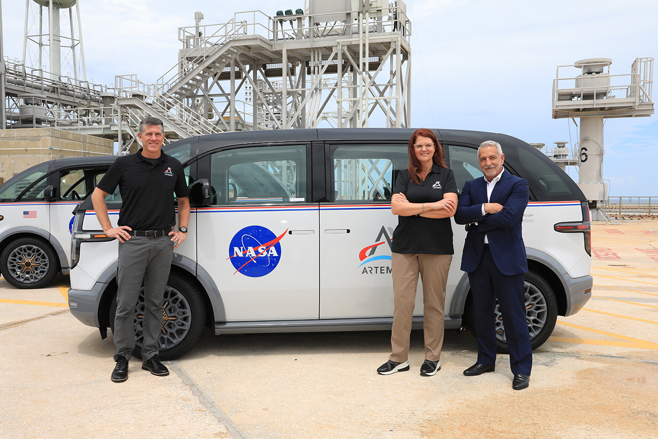 three people stand next to an electric minivan near a launch pad