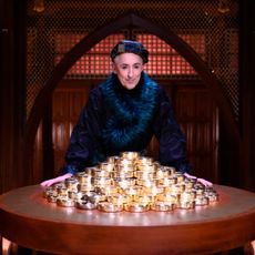 Host Alan Cumming leans on a round table with a pile of large gold coins, while standing in a chamber, in 'The Traitors' season 3.