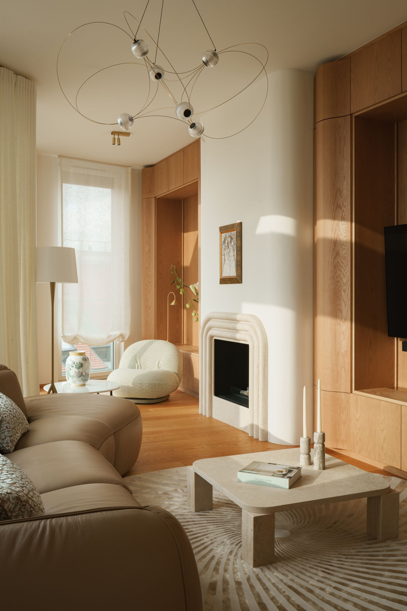Image of a modern living room that opens to a kitchen. The living room has wooden floors and walls, a beige leather bean-shaped sofa, a spiral rug, and a white marble coffee table.