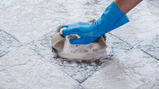 Hand with blue rubber glove holding a soapy sponge showing how to clean grout in floor tiles.