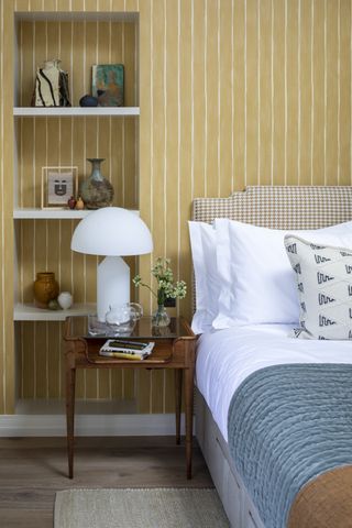 bedroom with yellow stripe walls, white bedding, white table lamp, blue throw, inset shelving