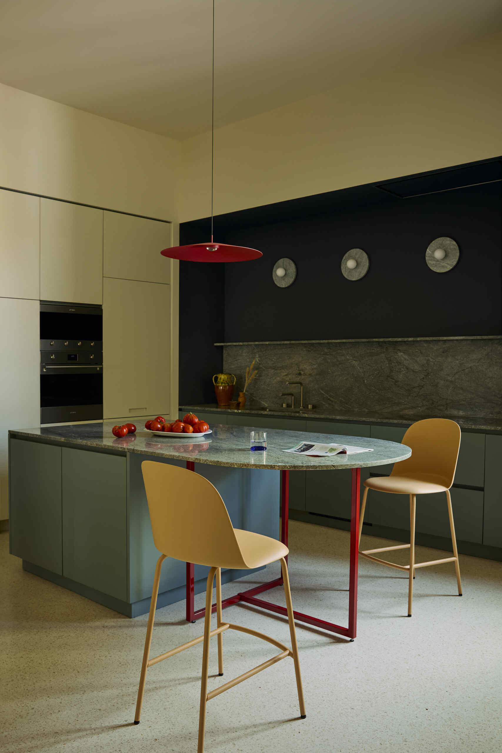 Image of a kitchen with off-white walls and white terrazzo flooring. There is a blue island, blue marble, and two orange bar stool chairs. The legs of the island are red and there is a red pendant.