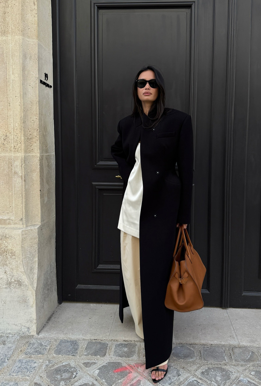 A woman standing on the sidewalk wearing a long black coat over a white satin blouse, ivory satin trousers, a brown bowler bag, black t-strap sandals, and black sunglasses.