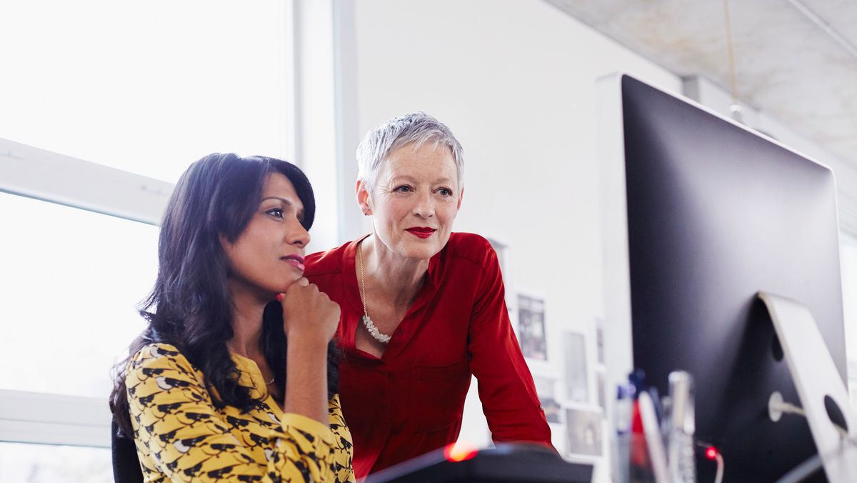 An older woman mentoring a younger colleague sat at a desk looking at a monitor.