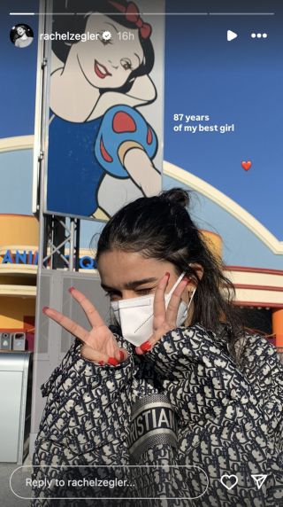 Rachel Zegler outside with a mask on holding up two peace signs in front of a Snow White vertical banner.