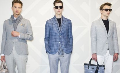 Three guys wearing the Hardy Amies S/S 2015 collection. On the left the guy is wearing a white shirt, gray scarf, checked gray jacket and gray pants. Next to him the guy is wearing a navy t-shirt, gray and navy striped scarf, blue checked jacket, gray pants and sunglasses. On the right the guy is wearing a navy t-shirt, gray jacket, white pants, sunglasses and holding a navy bag