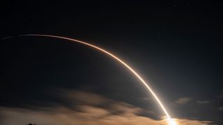 A rocket launch carves an orange arc into a dark night sky in this long-exposure photo.