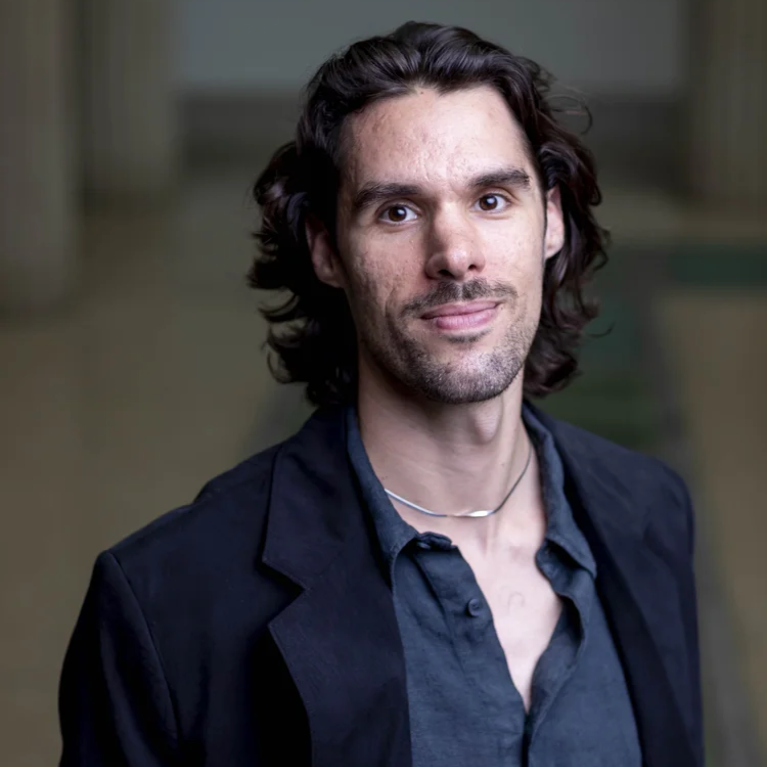 A headshot of a man with short wavy hair, a silver chain, a blue shirt, and a navy blazer