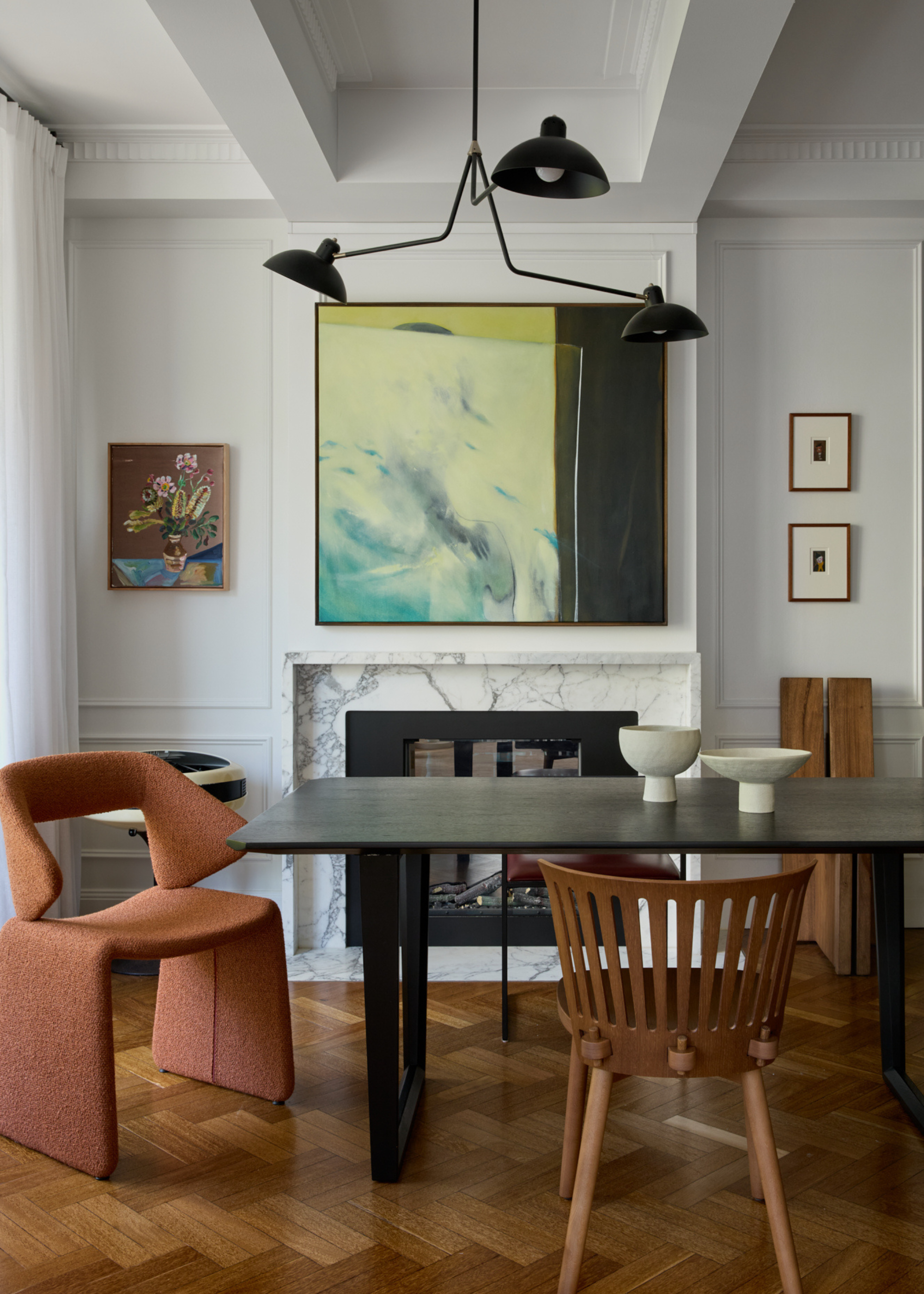 A living room with white walls, a stone fireplace, framed art on the walls, a metal black ceiling light, a dark wood dining table a set of white pedestal bowls, a wooden chair and a terracotta-colored sofa seat