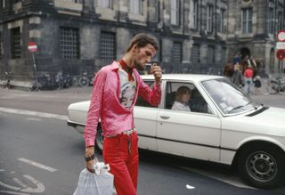 Dam, by Ed van der Elsken, 1975, daytime, street image, white car with female driving, no entry signs, stone building across the road, man wearing pink jacket, red trousers and white t-shirt, lighting a cigarette carrying a white plastic big, bicycles leant against the building, two adults and small child gathered near mounted police horses in the distance