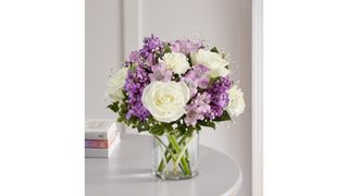 A medium bouquet of white roses and carnations, purple lilies, and assorted greenery in a clear cylinder vase, for the best flower delivery services.