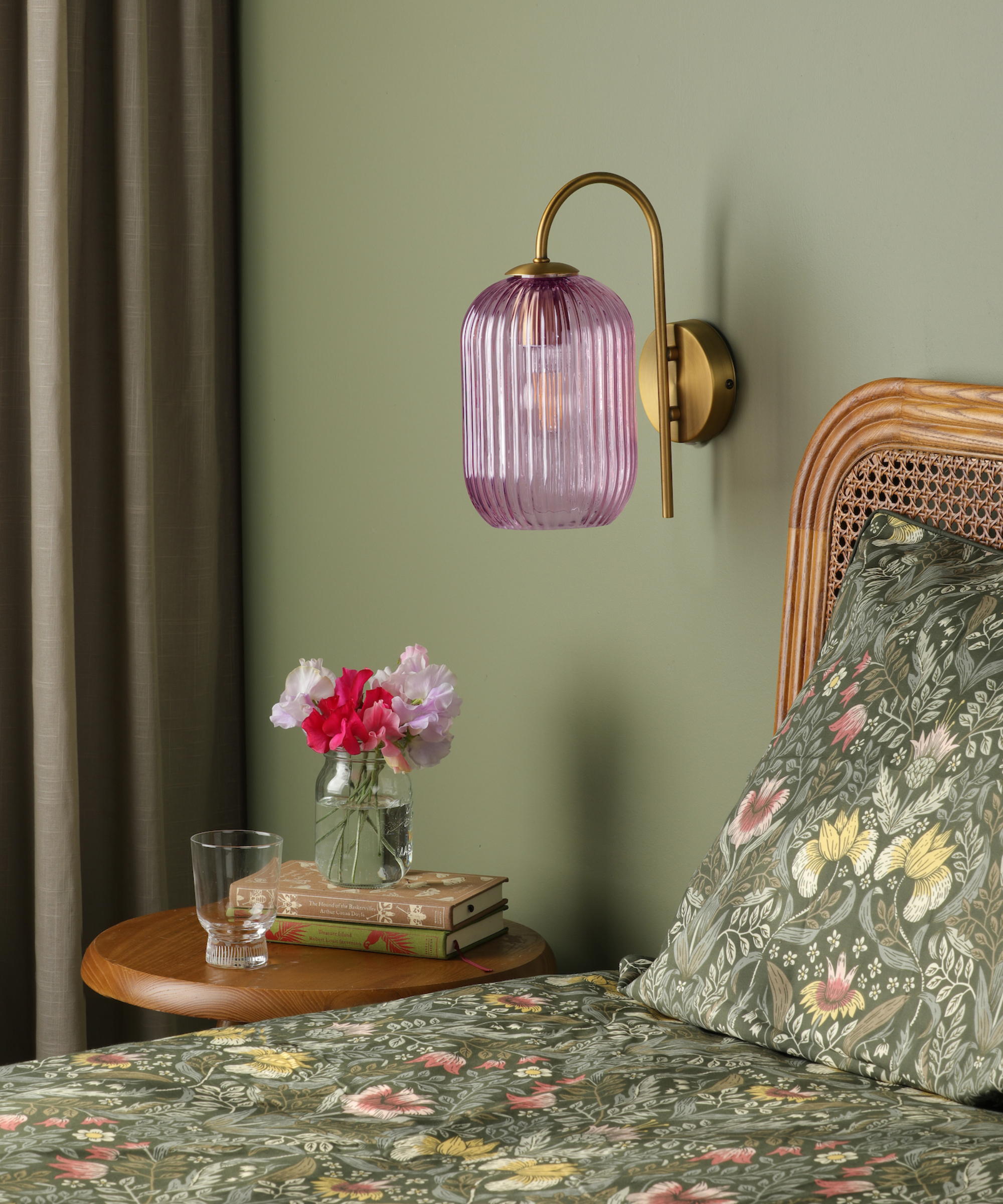 gold wall lamp with pink shade on green painted bedroom wall with rattan bedframe and small round wooden table to side of bed