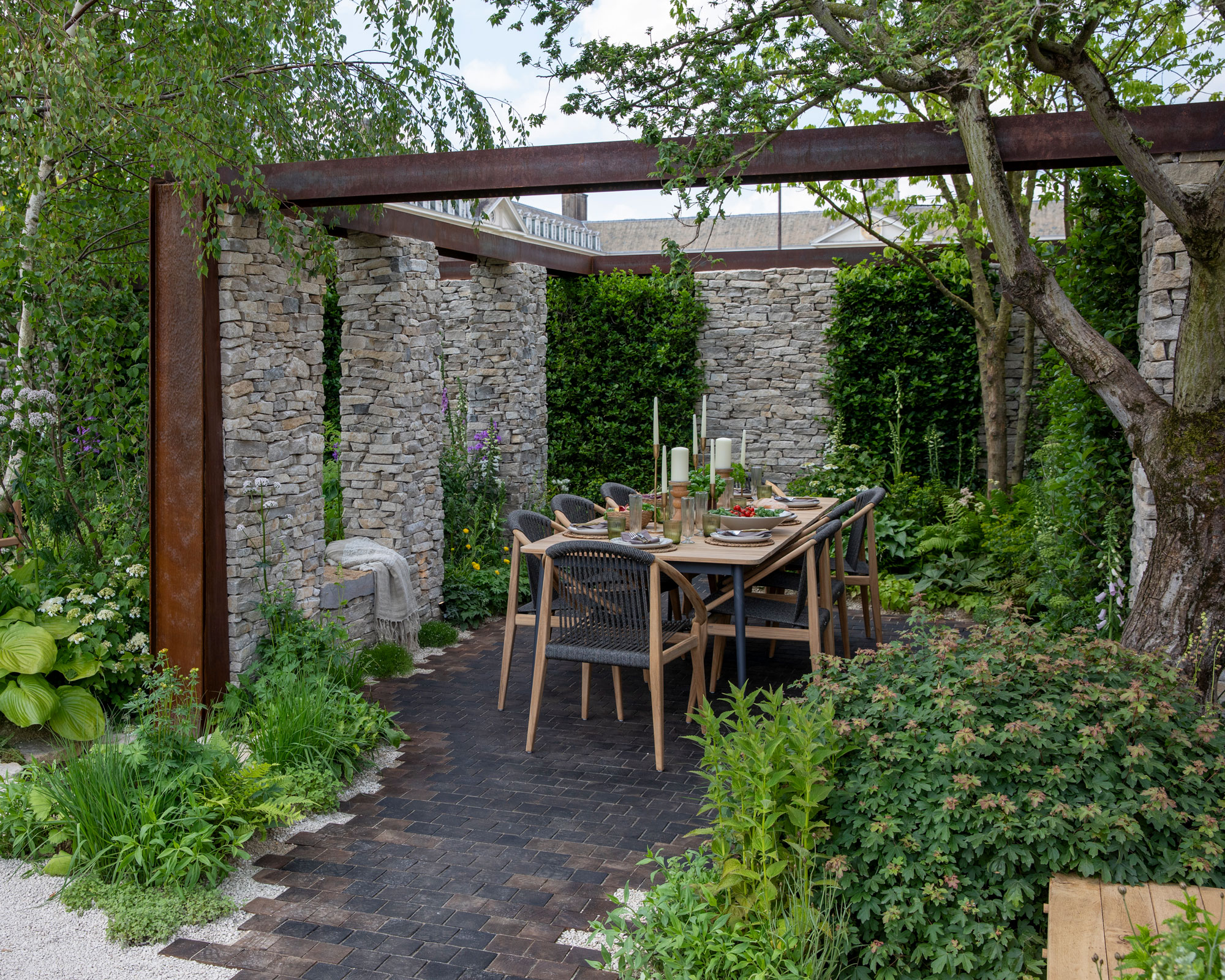 pergola design filled in with stone wall and living wall sections, outdoor dining table and chairs, brick patio, and planting including trees and shrubs