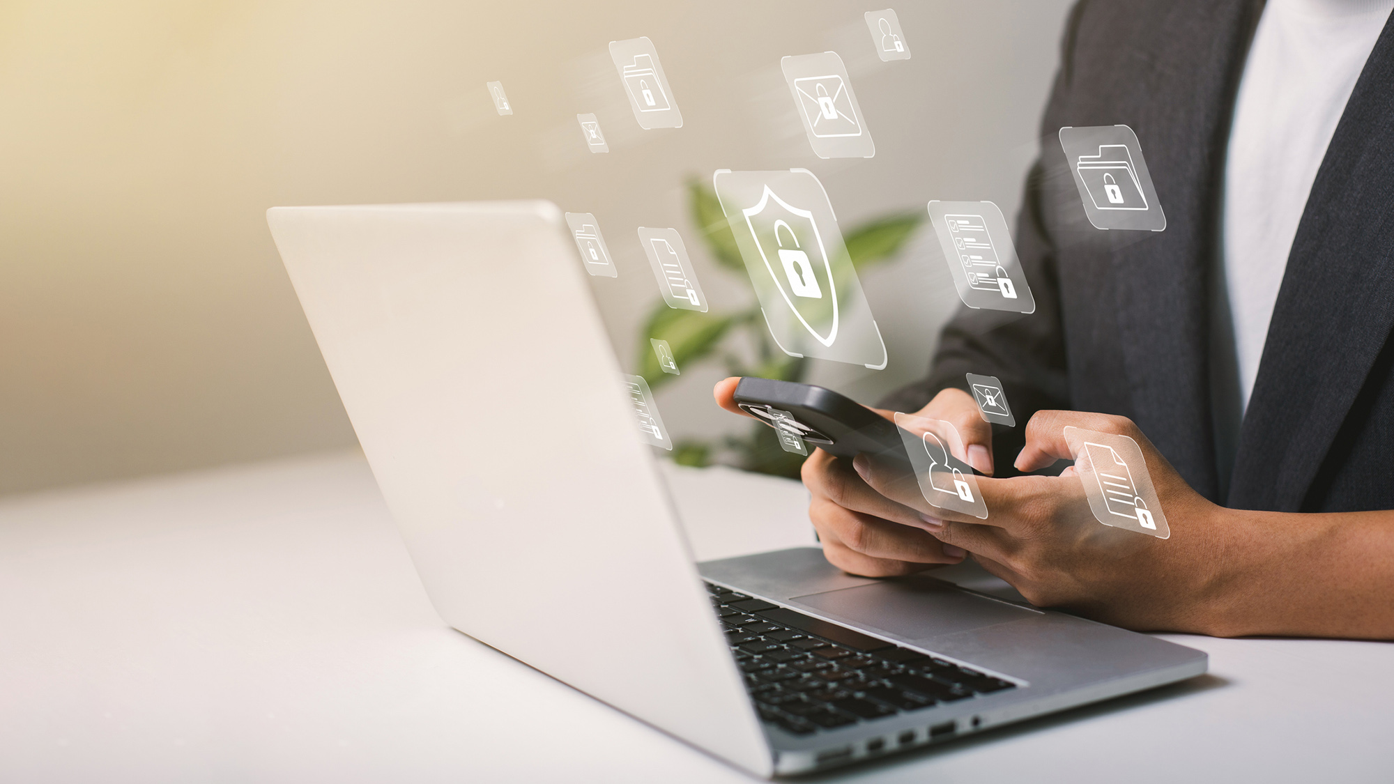 A person sitting at a laptop holding in their hands a smartphone with icons such as a padlock in a shield and a file with a padlock on it floating around it. Their face is not visible.