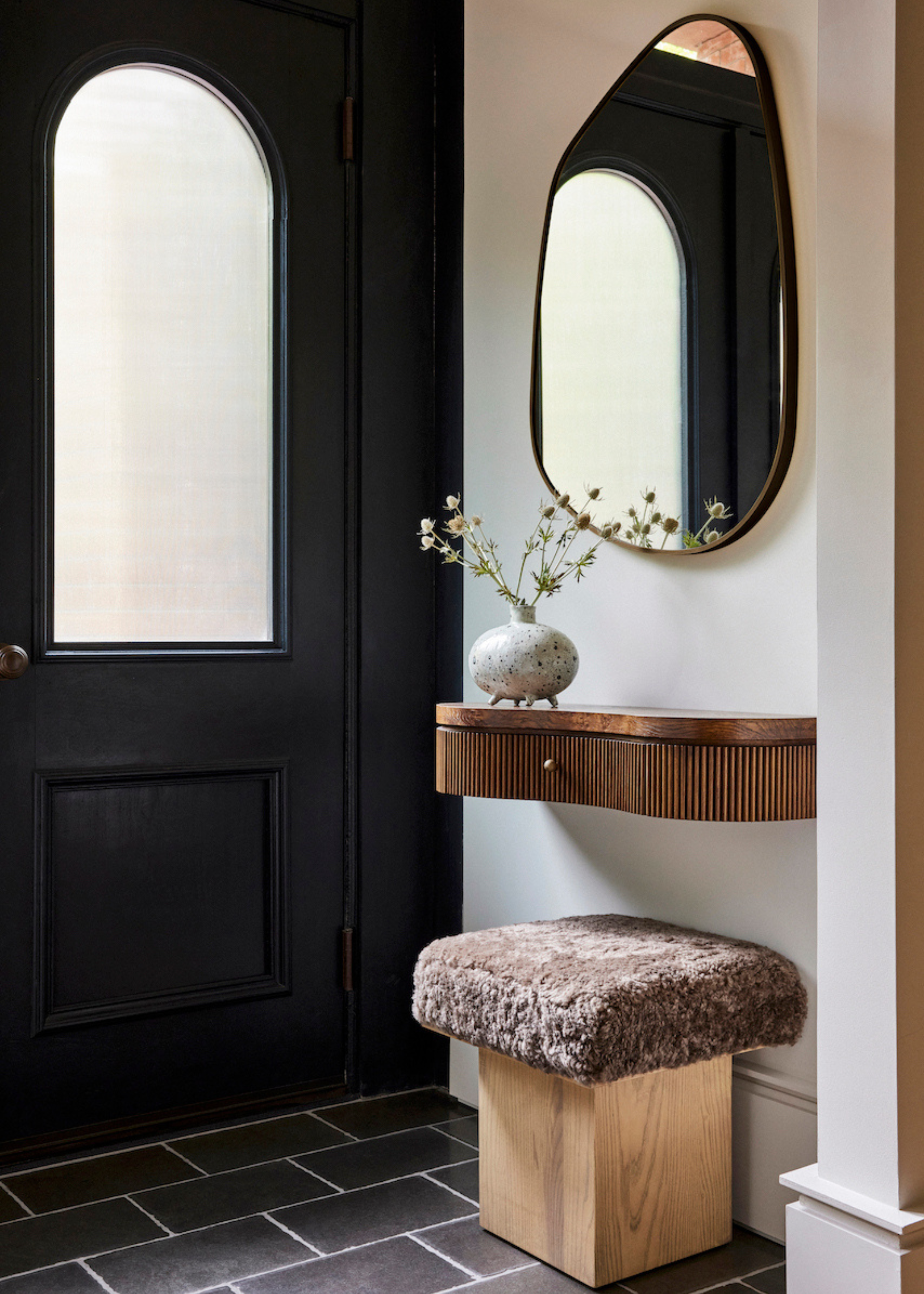 An entryway with a hanging mirror, a floating table and a fuzzy stool by a black door
