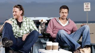 (L, R) DONAL LOGUE and MICHAEL RAYMOND-JAMES sit in the back of a pickup truck with a barking dog in the art for Terriers Terriers