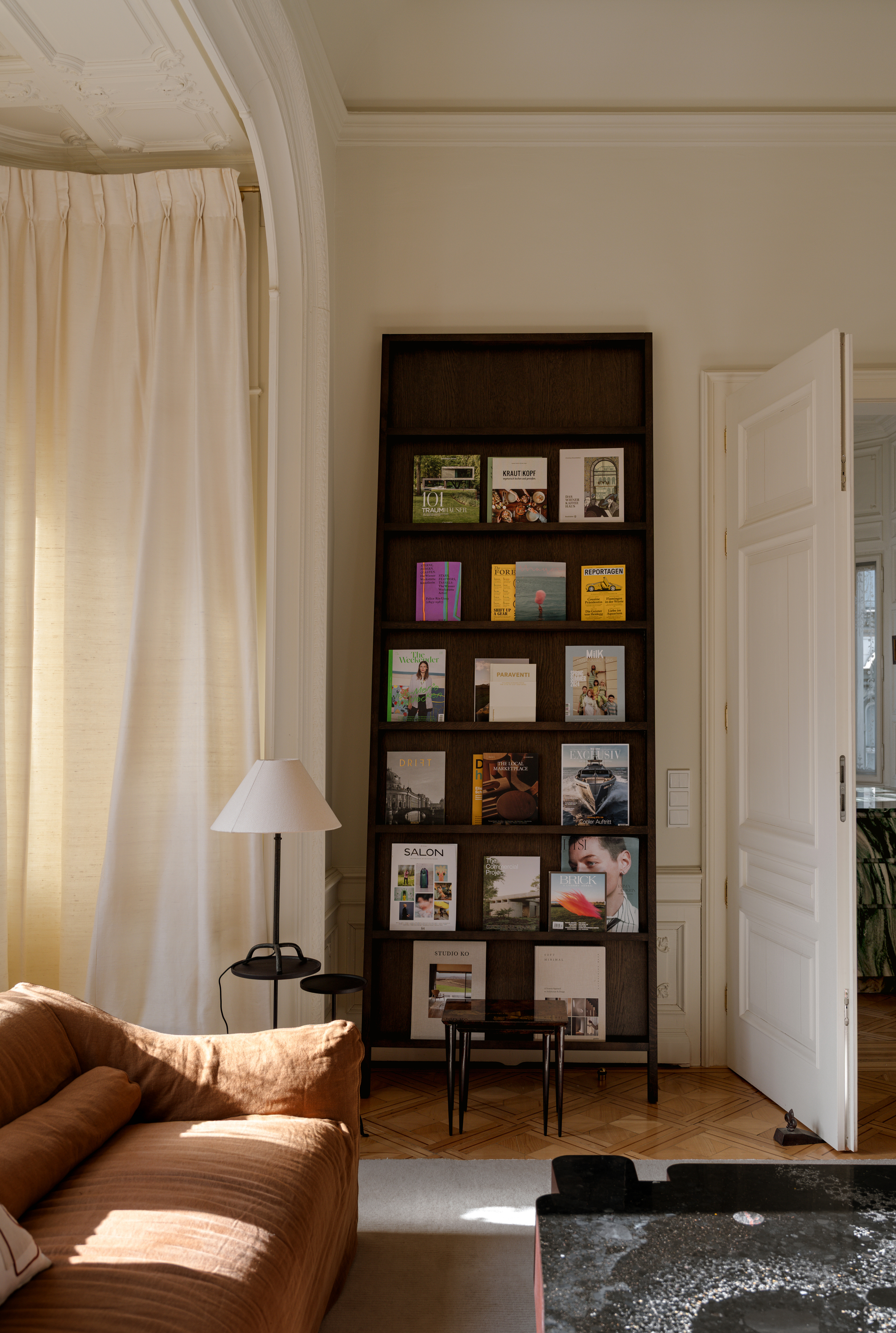 Image of a large dark brown wooden magazine stand leaning against a white wall in a grand living room. There is a side table with a lamp on it, a brown sofa, and a bay window with white curtains in view.
