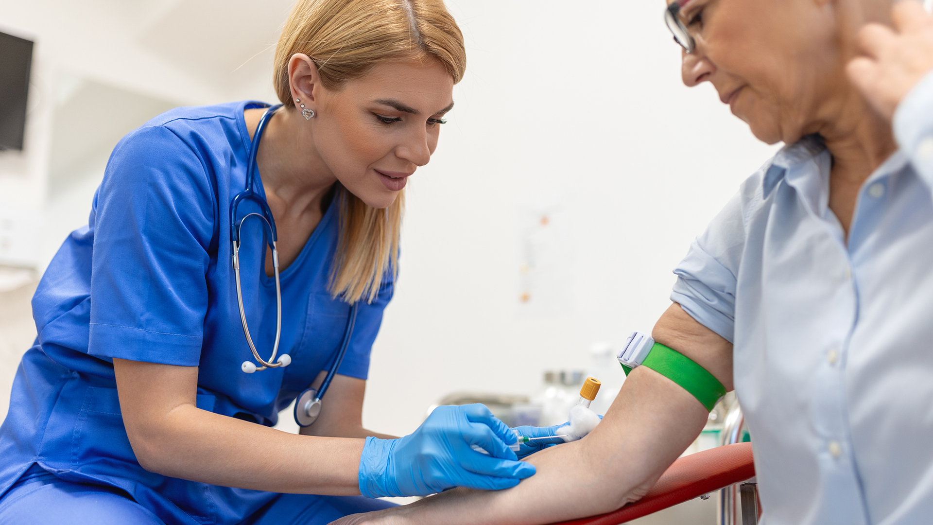A blond woman with blue scrubs pokes a needle into the vein of a person with glasses and a button-down shirt